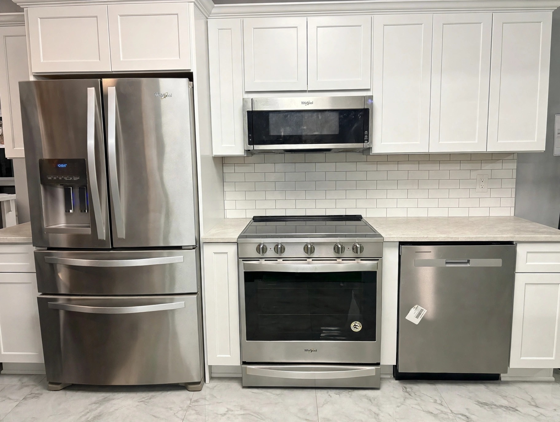 Whirlpool 4-piece stainless kitchen package installed in a showroom kitchen display — French-door refrigerator, slide-in range with over-range microwave, and dishwasher